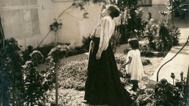 Num jardim, mãe e filha estão em pé e são vistas de lado. A mãe olha para cima, a filha carrega um cesto.