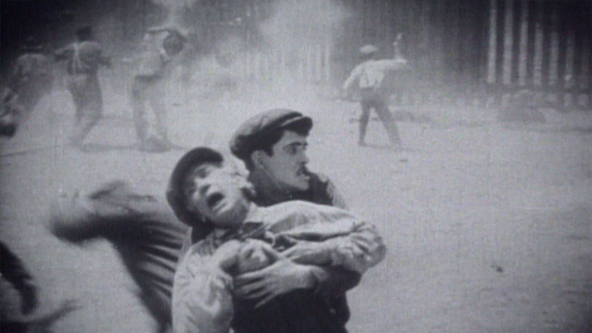 Na frente, um homem segura outro que grita. No fundo, outros homens em meio à fumaça. Cena de filme antigo.