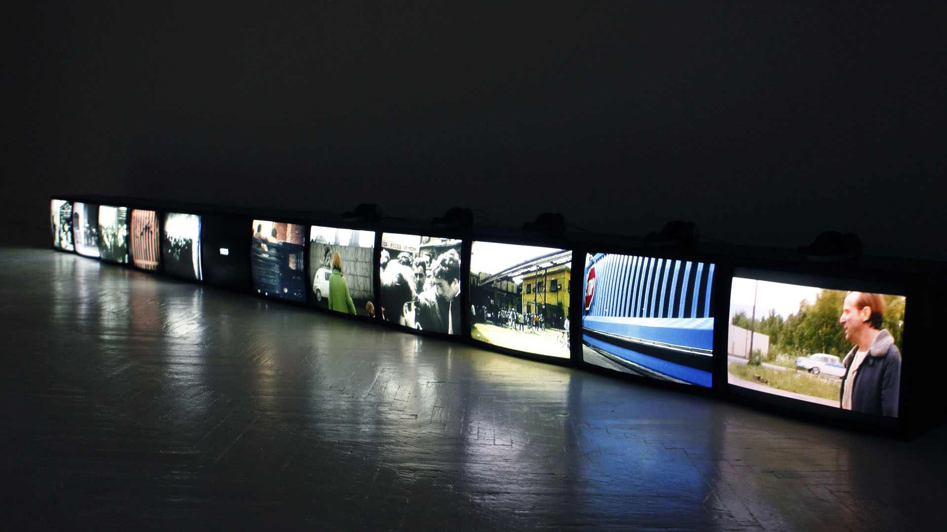 Doze telas de vídeo, lado a lado no chão, exibem cenas da obra de Harun Farocki