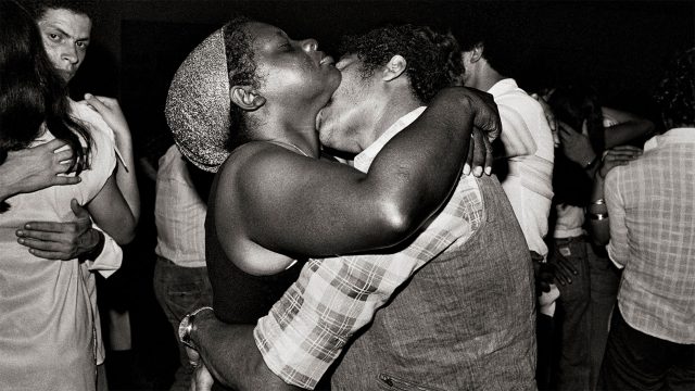 Fotografia preto e branco. No centro: casal dançando forró. O homem beija o pescoço da mulher, que está de olhos fechados. Ao fundo, outros casais dançam. À esquerda, um homem olha para a câmera.