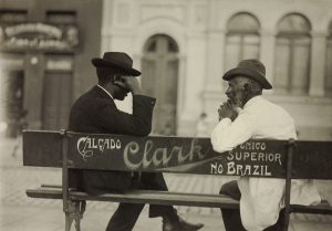 Os dois homens estão sentados e aparecem de perfil. Estão de terno e chapéu. O banco é visto por trás e tem uma propaganda de "Calçado Clark" no encosto.
