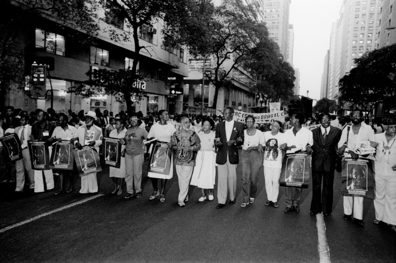 Marcha Zumbi está Vivo. Av. Rio Branco. Rio de Janeiro, 18/11/1983. Na imagem, da esquerda para a direita: 1. Alberto Vieira; 2. não identificado; 3. Eduardo de Oliveira; 4. Roberto Ananias (Cordão Carnavalesco, Educativo e Cultural, de Samba, Frevo e Capoeira “Divina Senzala” - SAMFRECAPO D.S.); 5. Dr.ª Sebastiana Arruda (Irmandade Nossa Senhora do Rosário São Benedito dos Homens dos Pretos); 6. Lélia Gonzalez; 7. Benedita da Silva (então, vereadora pelo PT); 8. Abdias do Nascimento; 9. Dr.ª Edialeda Salgado do Nascimento; 10. José Miguel; 11. não identificada; 12. Lili Paiva (Rocinha); 13. não identificado; 14. não identificado; 15. não identificado; 16. Sebastião Rodrigues Alves. Foto de Januário Garcia. Acervo IMS /Arquivo Januário Garcia