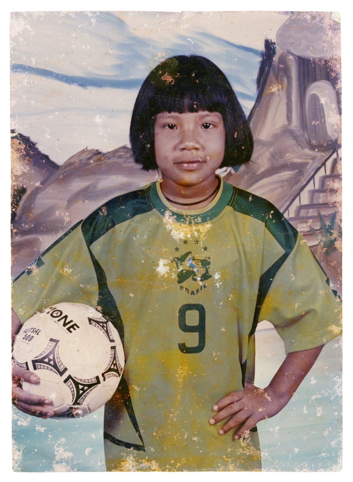 Rebeca kaban Waleguere Suru&iacute;, Cacoal, RO, anos 1990 O futebol &eacute; de longe o esporte mais amado e praticado na Terra Ind&iacute;gena Sete de Setembro. Na d&eacute;cada de 1990 o Brasil n&atilde;o s&oacute; teve muitas vit&oacute;rias, mas tamb&eacute;m equipes repletas de estrelas. A "amarelinha" era muito popular entre os Paiter. Ap&oacute;s o tetracampeonato de 1994, a m&atilde;e de Rebeca a levou em um est&uacute;dio na cidade para fazer este retrato. S&atilde;o poucas as fotos que restaram de sua inf&acirc;ncia, ent&atilde;o esta lembran&ccedil;a ganhou ainda mais import&acirc;ncia para a fam&iacute;lia. Autoria desconhecida. (Legenda por Coletivo Lakapoy).  Fotografias em pigmento mineral sobre papel de algod&atilde;o impressas em 2025. 52 x 70,3 cm. Cole&ccedil;&atilde;o de Arte Contempor&acirc;nea / Acervo IMS 