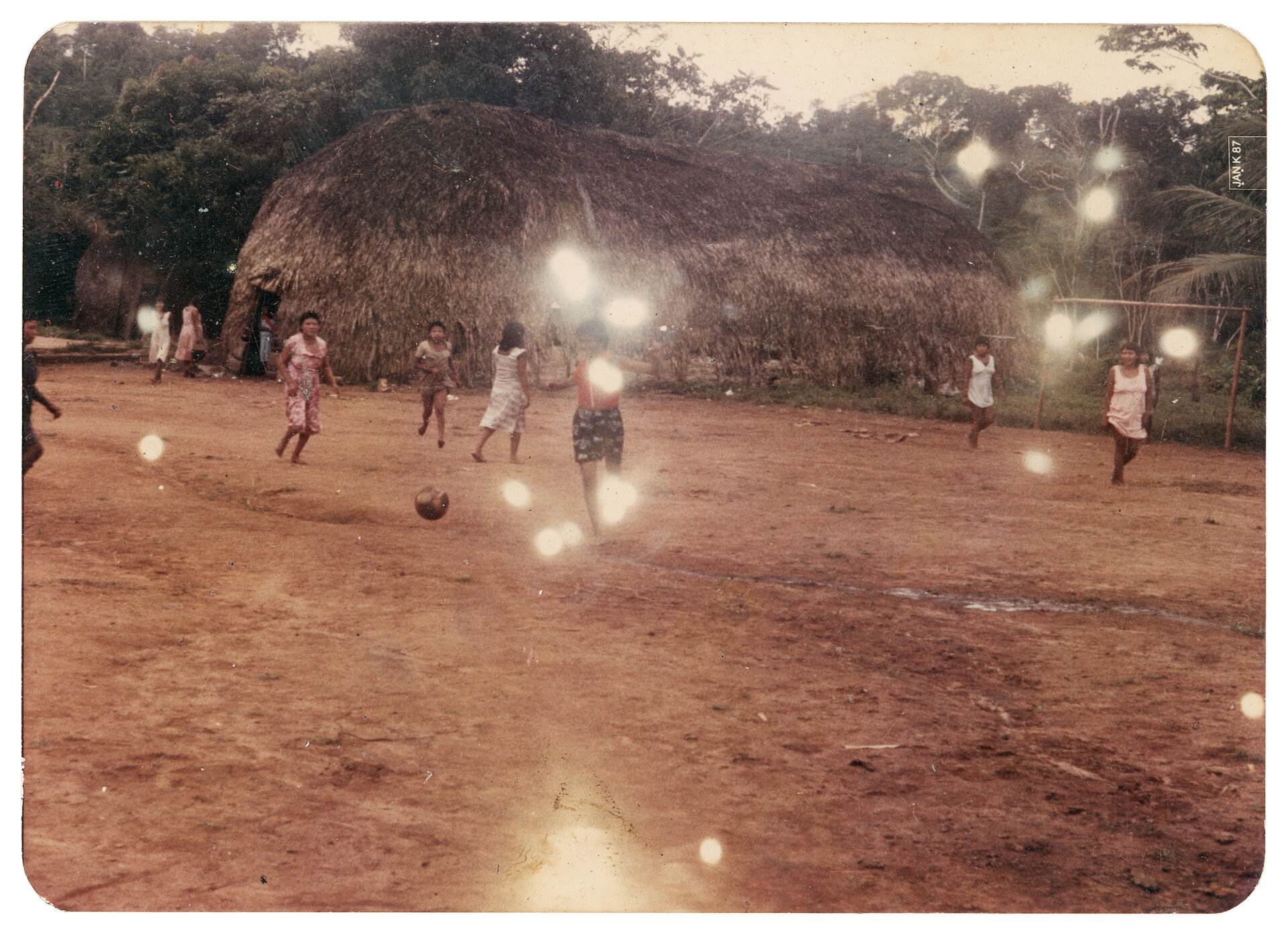 Jogo de futebol feminino, Aldeia Lapetanha, c. 1986 As partidas de futebol entre as aldeias Lapetanha e Joaquim marcaram &eacute;poca. As mulheres jogavam equipadas apenas com vestidos fornecidos pela Funai. Esses jogos, que tamb&eacute;m envolviam outros cl&atilde;s, davam pr&ecirc;mios para os vencedores, que inclu&iacute;am artesanatos, carne de ca&ccedil;a e outros itens tradicionais, transformando-se em um importante momento de integra&ccedil;&atilde;o entre as comunidades. A rivalidade saud&aacute;vel e a troca de presentes criavam um clima de festa e uni&atilde;o, fortalecendo os la&ccedil;os entre os diferentes grupos e n&uacute;cleos familiares. Foto da missionaria Tine Wand Mer. (Legenda por Coletivo Lakapoy).  Fotografias em pigmento mineral sobre papel de algod&atilde;o impressas em 2025. 92 x 67,3 cm. Cole&ccedil;&atilde;o de Arte Contempor&acirc;nea / Acervo IMS   